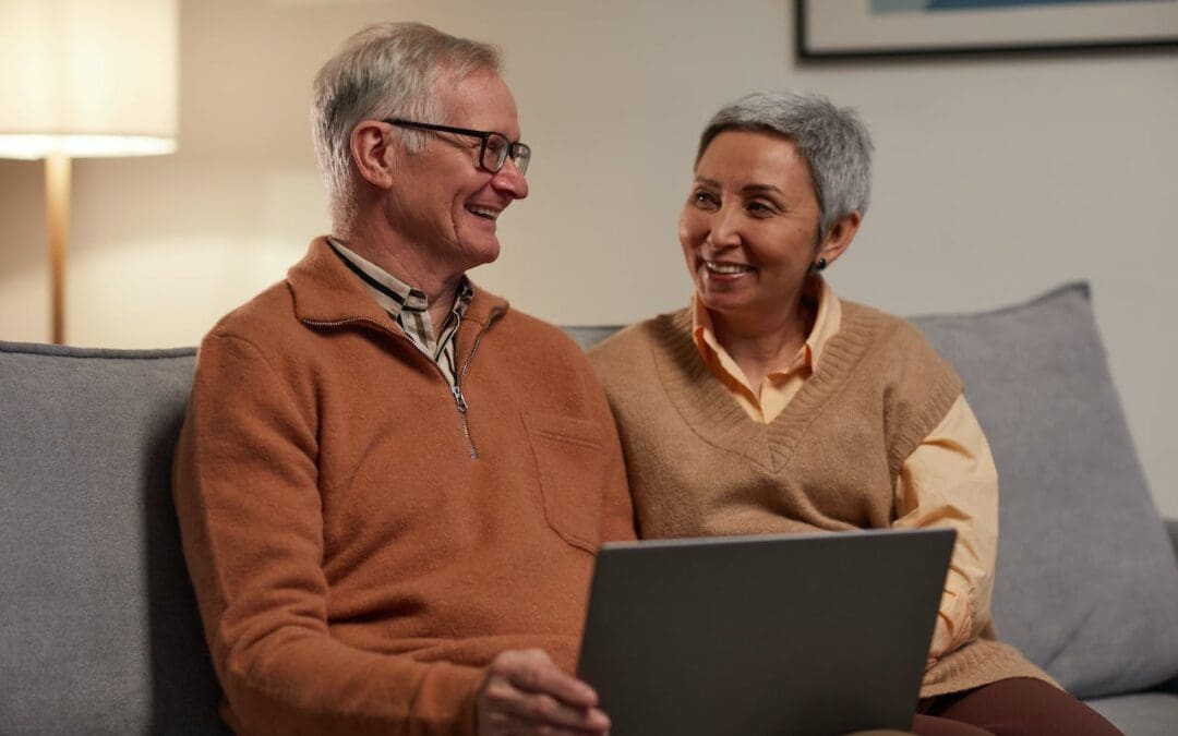 A couple in their 50s sit on a couch and smile at each other. The man, who wears glasses, holds a computer in his lap.